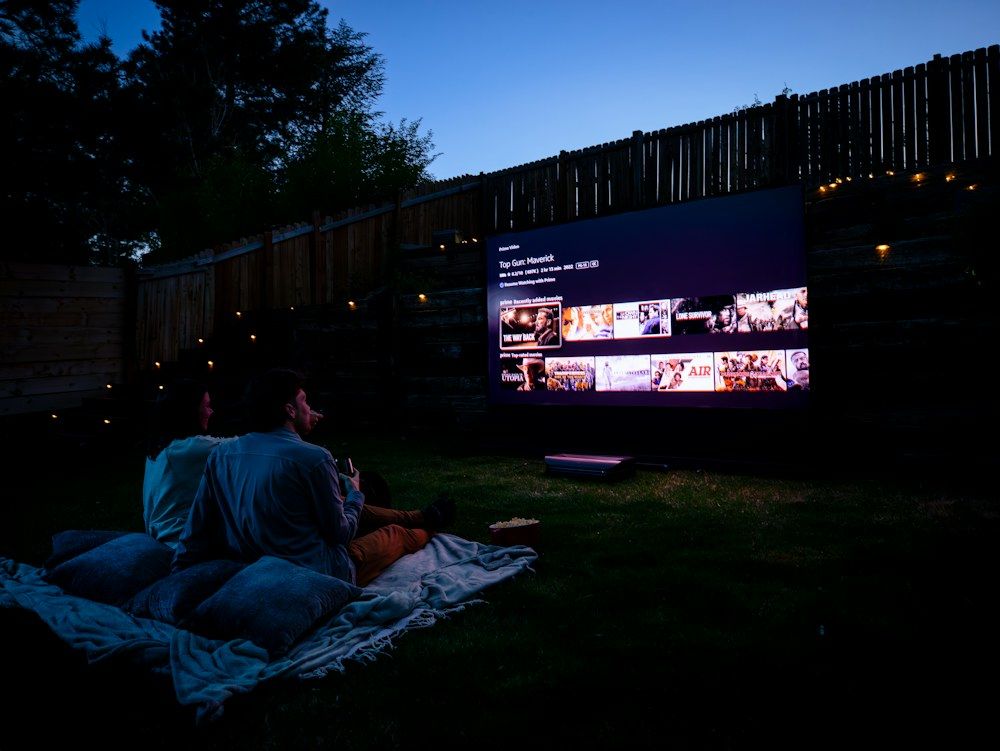 A cozy backyard scene unfolds as two people sit on a blanket, immersed in an outdoor cinema experience.