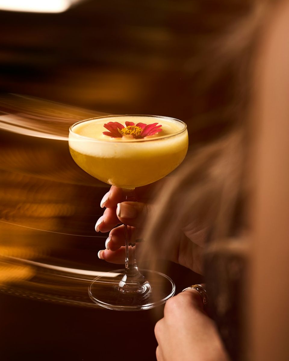 A coupe glass brimming with a frothy yellow cocktail is held by a hand, its surface adorned with a single red flower.
