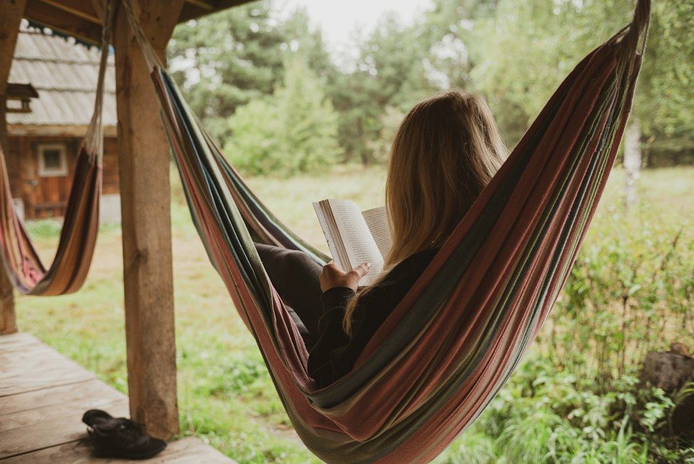 A young woman sits comfortably in a striped hammock tied to the beams of a wooden porch, engrossed in her book.